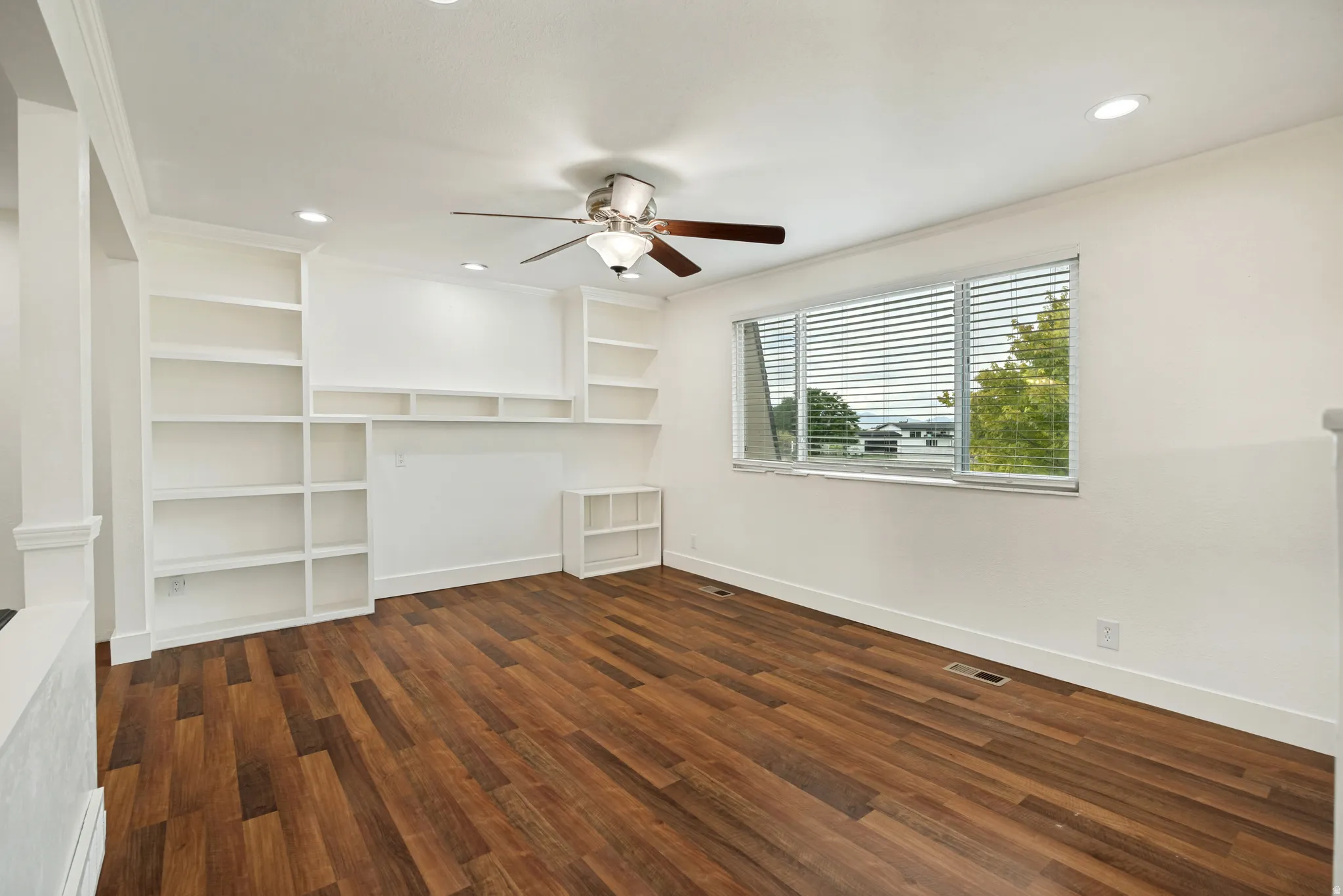 Wood-finish flooring, recessed lighting, ceiling fan, built-in shelving, and a large window with blinds