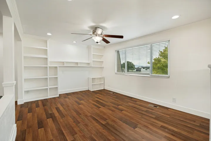 Wood-finish flooring, recessed lighting, ceiling fan, built-in shelving, and a large window with blinds