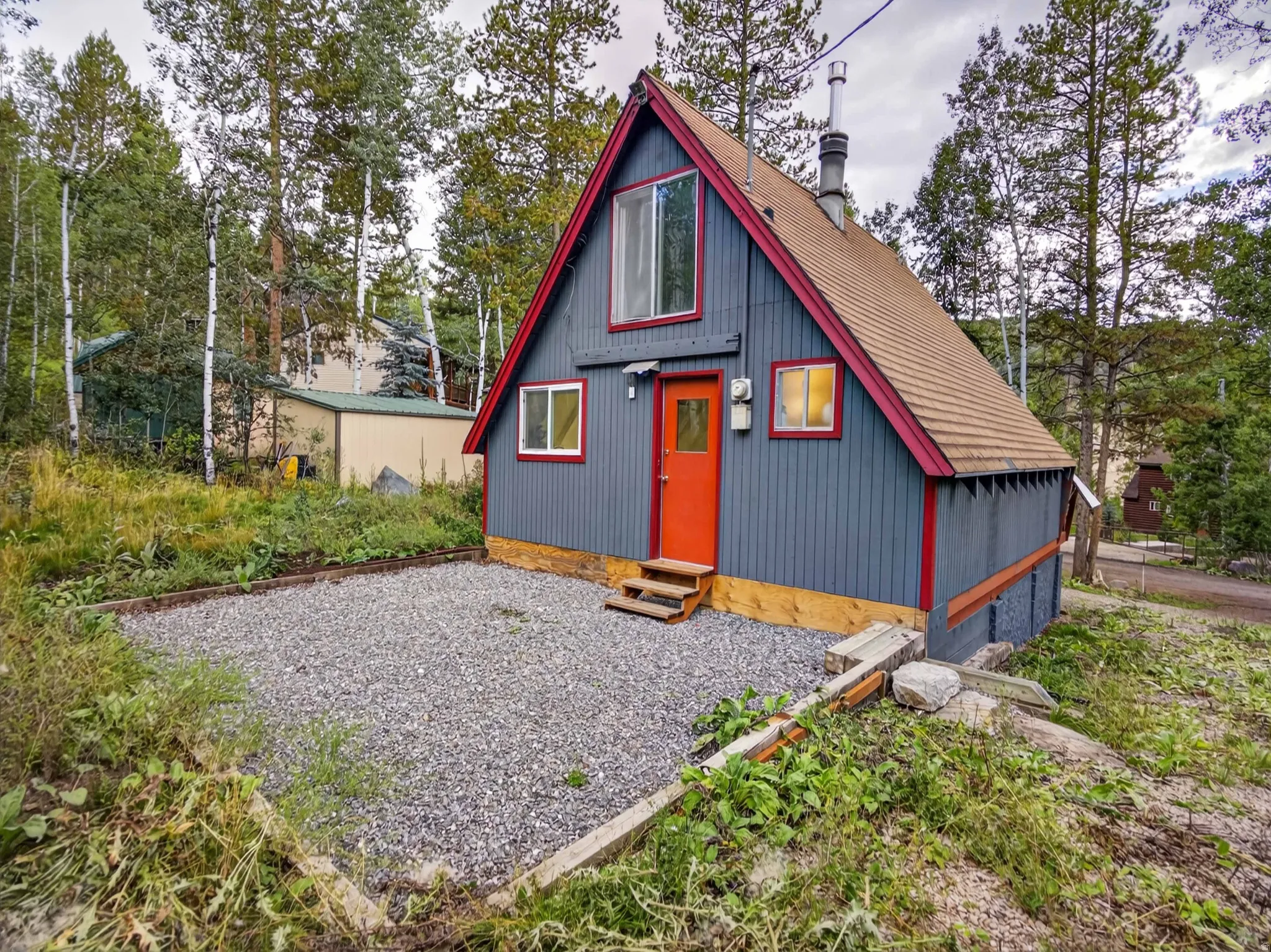 A-frame structure with gray siding, a prominent red front door, a light-colored shingle roof, and a gravel ground cover