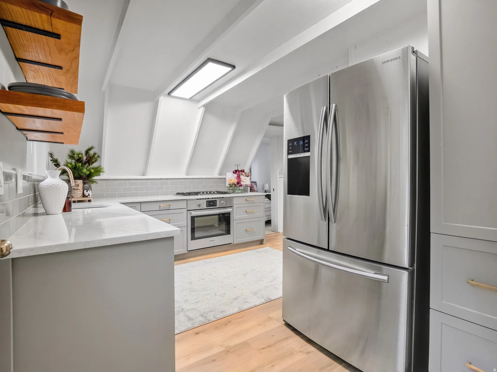 Kitchen featuring light wood-finish flooring, light gray cabinetry with gold-tone hardware, white countertops, stainless steel appliances, and a tiled backsplash