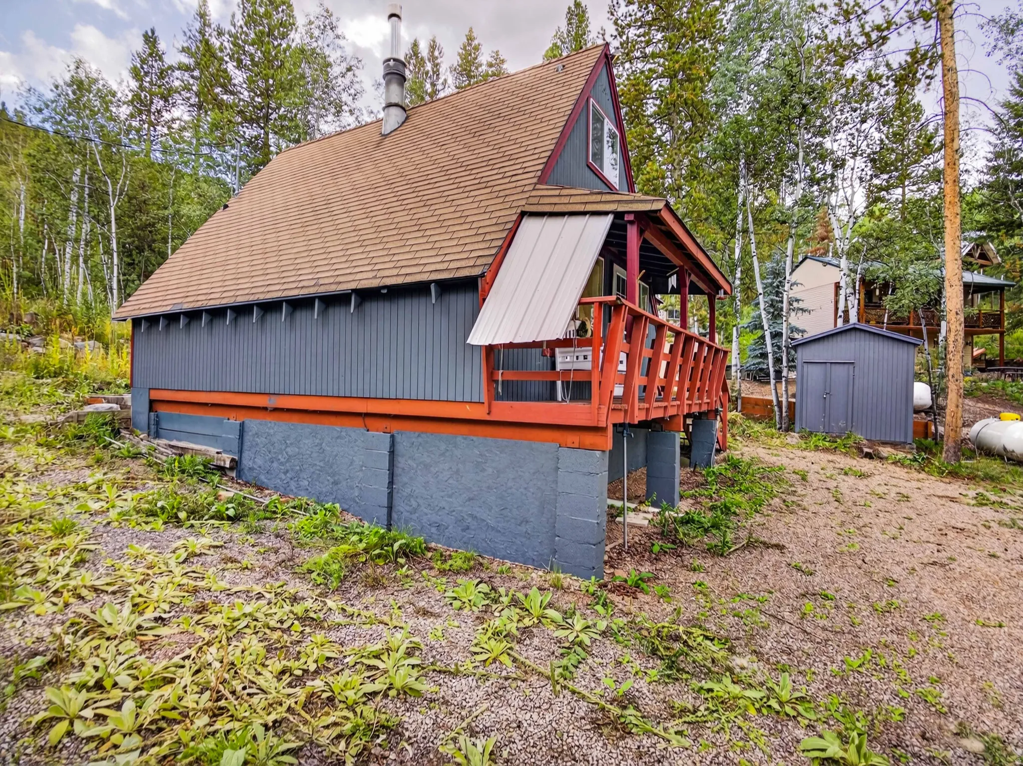 A-frame structure with a pitched roof, featuring a raised deck with wood railings, a dedicated utility shed, and a prominent chimney