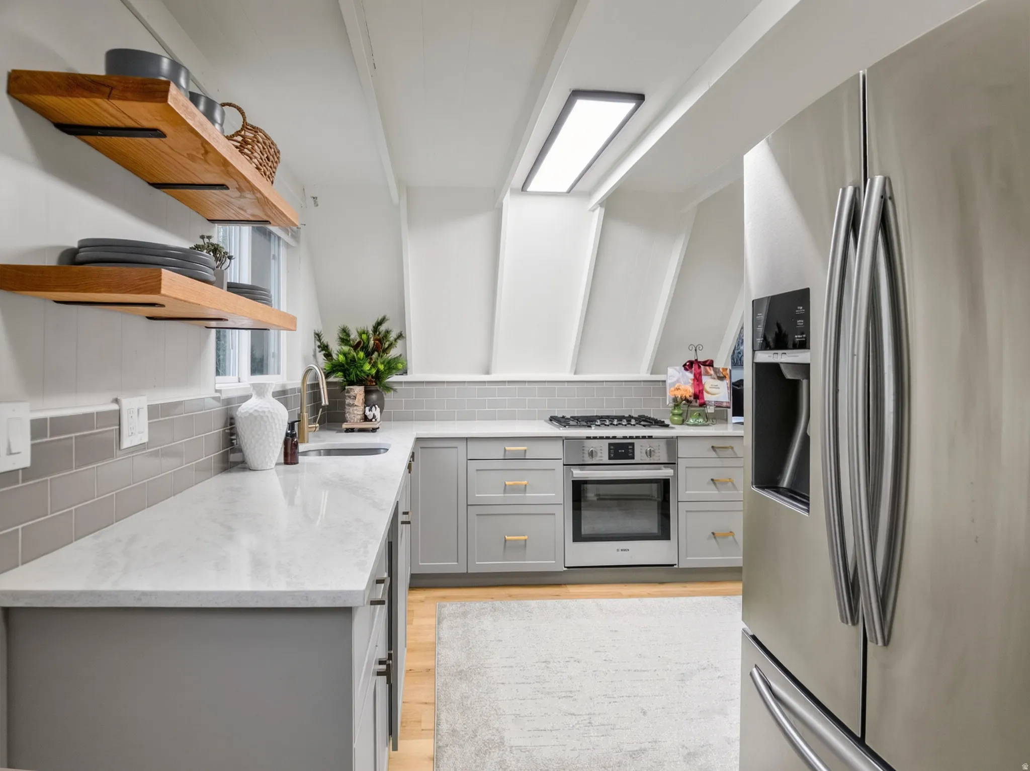 Light gray cabinetry with gold-tone hardware, an undermount sink with a gooseneck faucet, light-toned countertops, and a subway tile backsplash