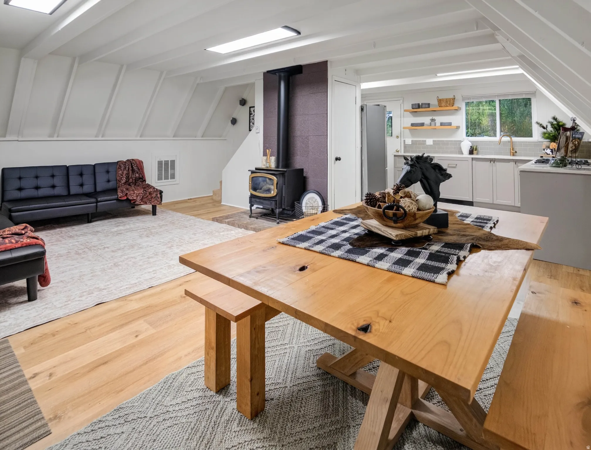 Vaulted ceiling with exposed beams, wood-finish flooring, wood-burning stove with a dark tile surround, kitchen with white cabinetry and open shelving, and a dual basin sink with a gold-tone faucet