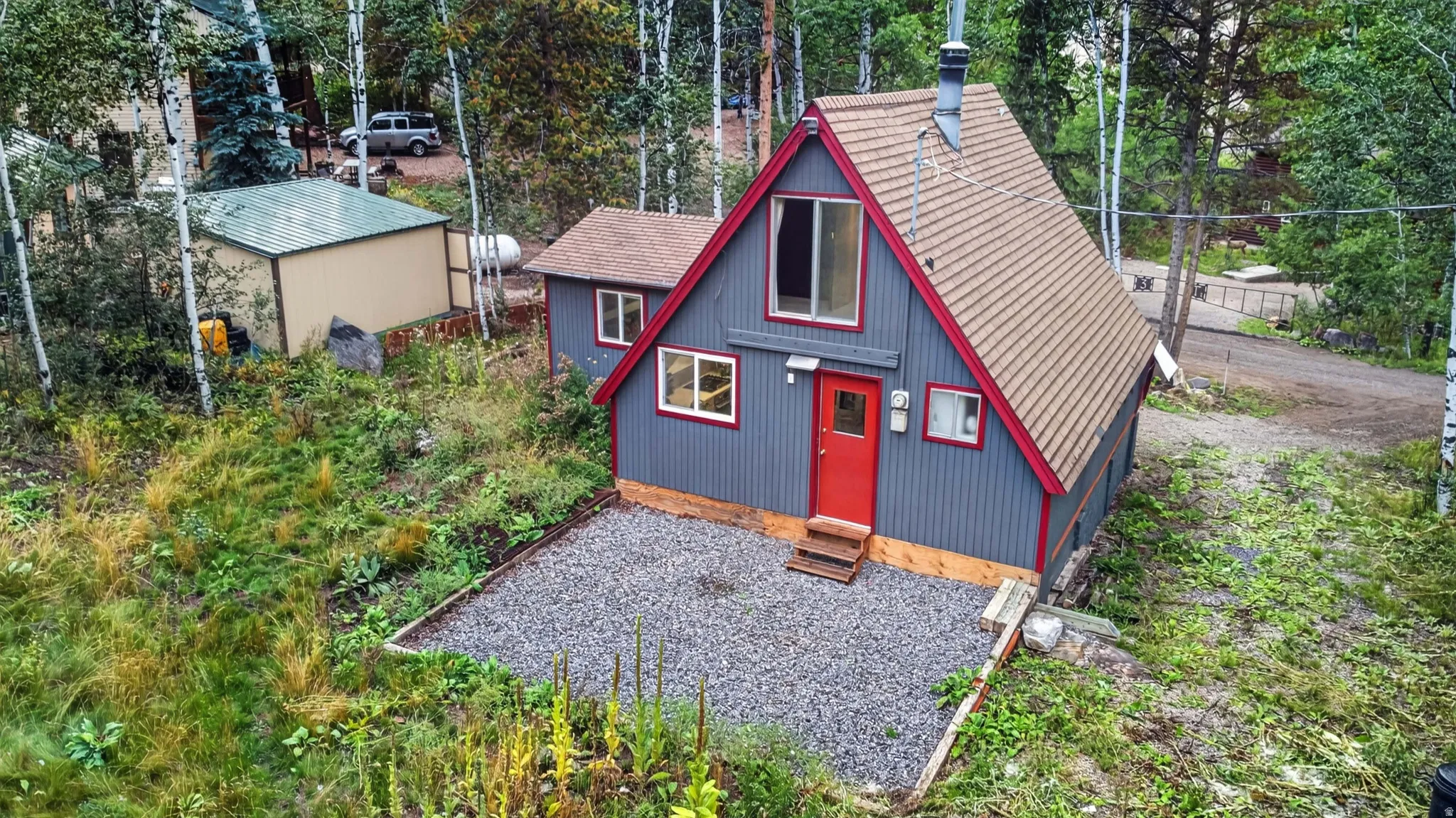 A-frame style residence with red trim accents, a vibrant red entry door, a gravel parking area, and an exterior storage shed