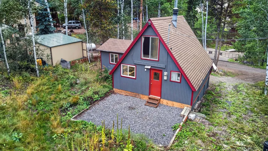 A-frame style residence with red trim accents, a vibrant red entry door, a gravel parking area, and an exterior storage shed