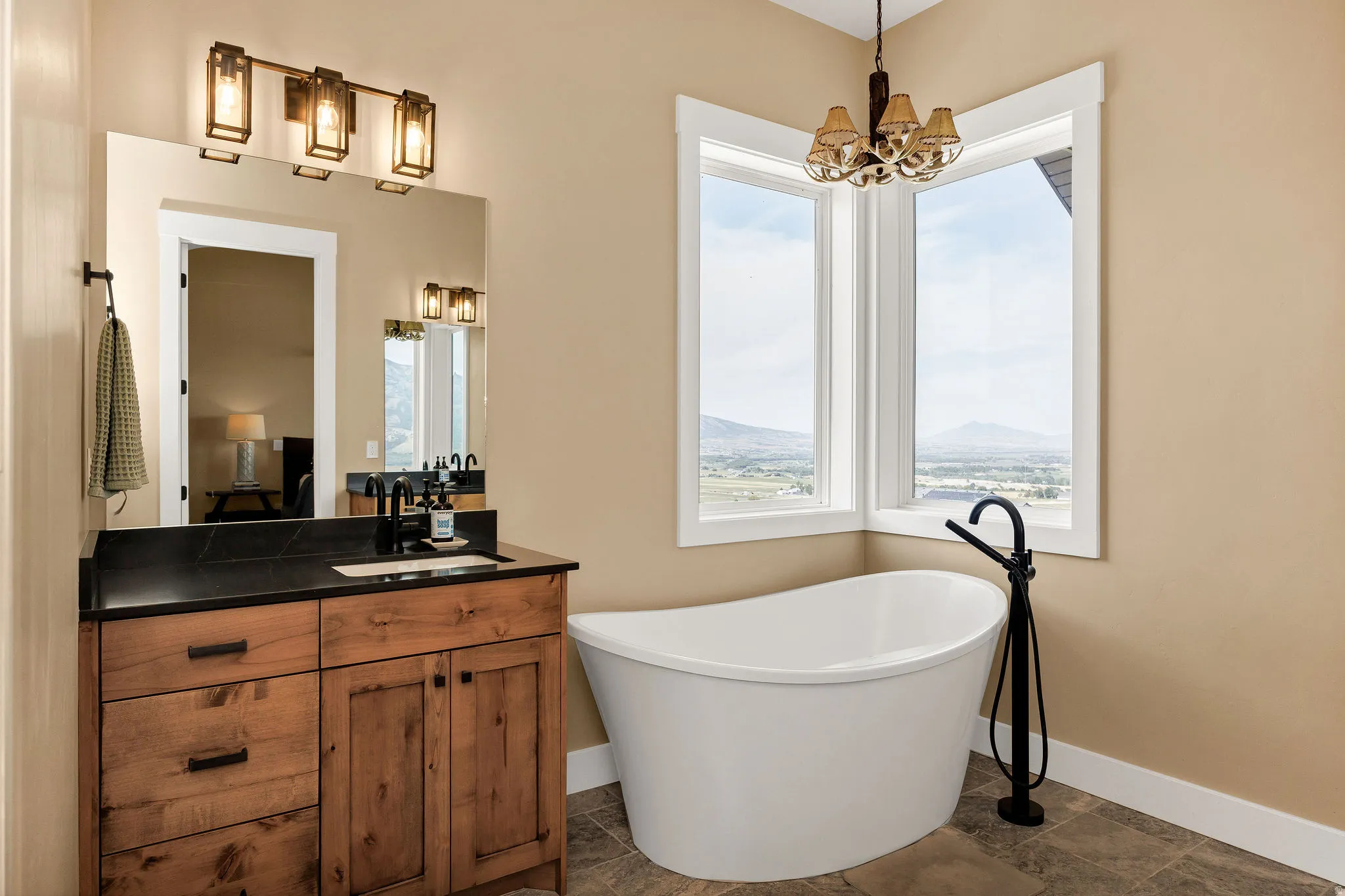 Freestanding soaking tub with floor-mounted fixture, wood-finish vanity with dark countertop, large vanity mirror, and corner windows