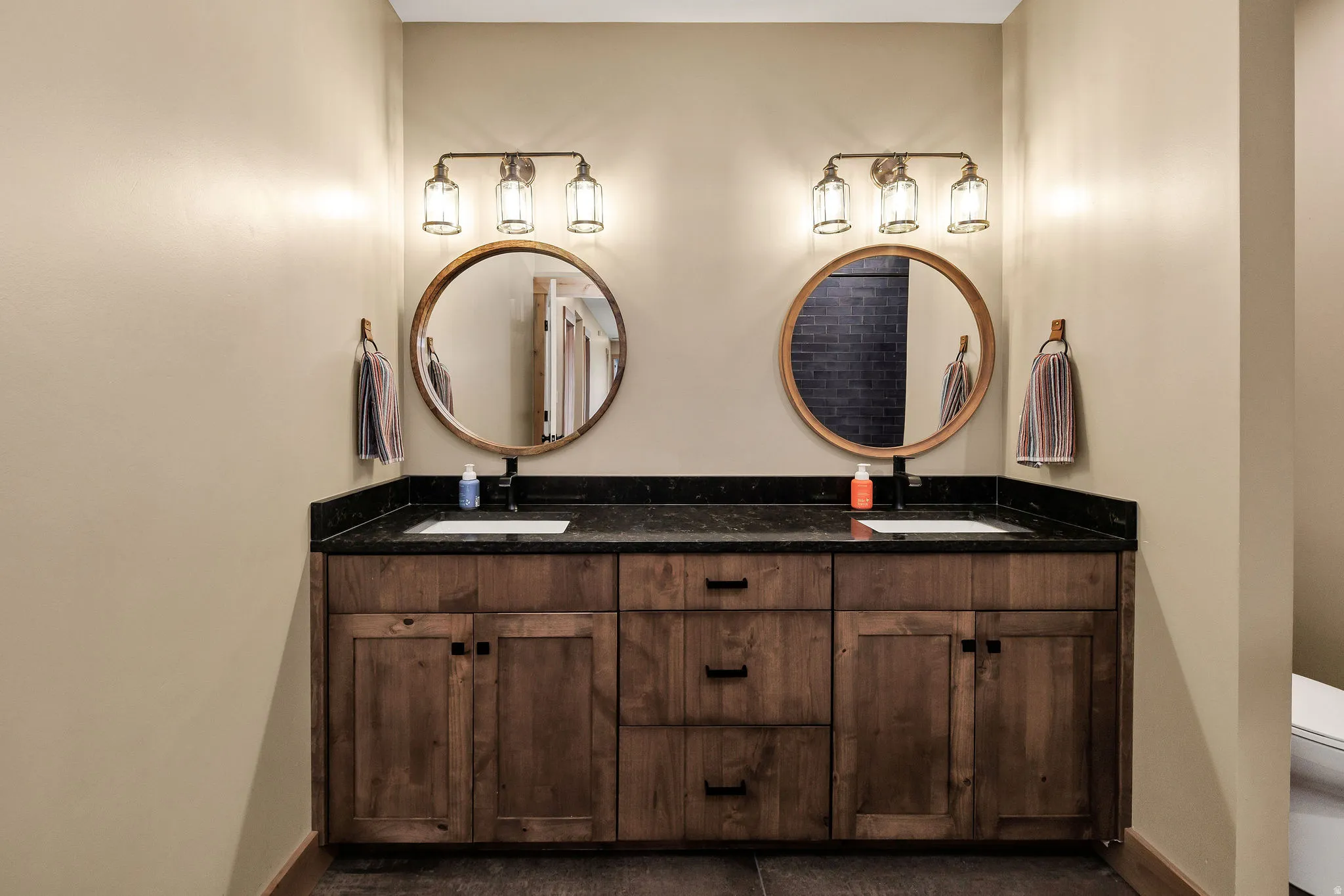 Dual wood-finish vanities with black countertops, integrated sinks, and individual round mirrors with sconce lighting