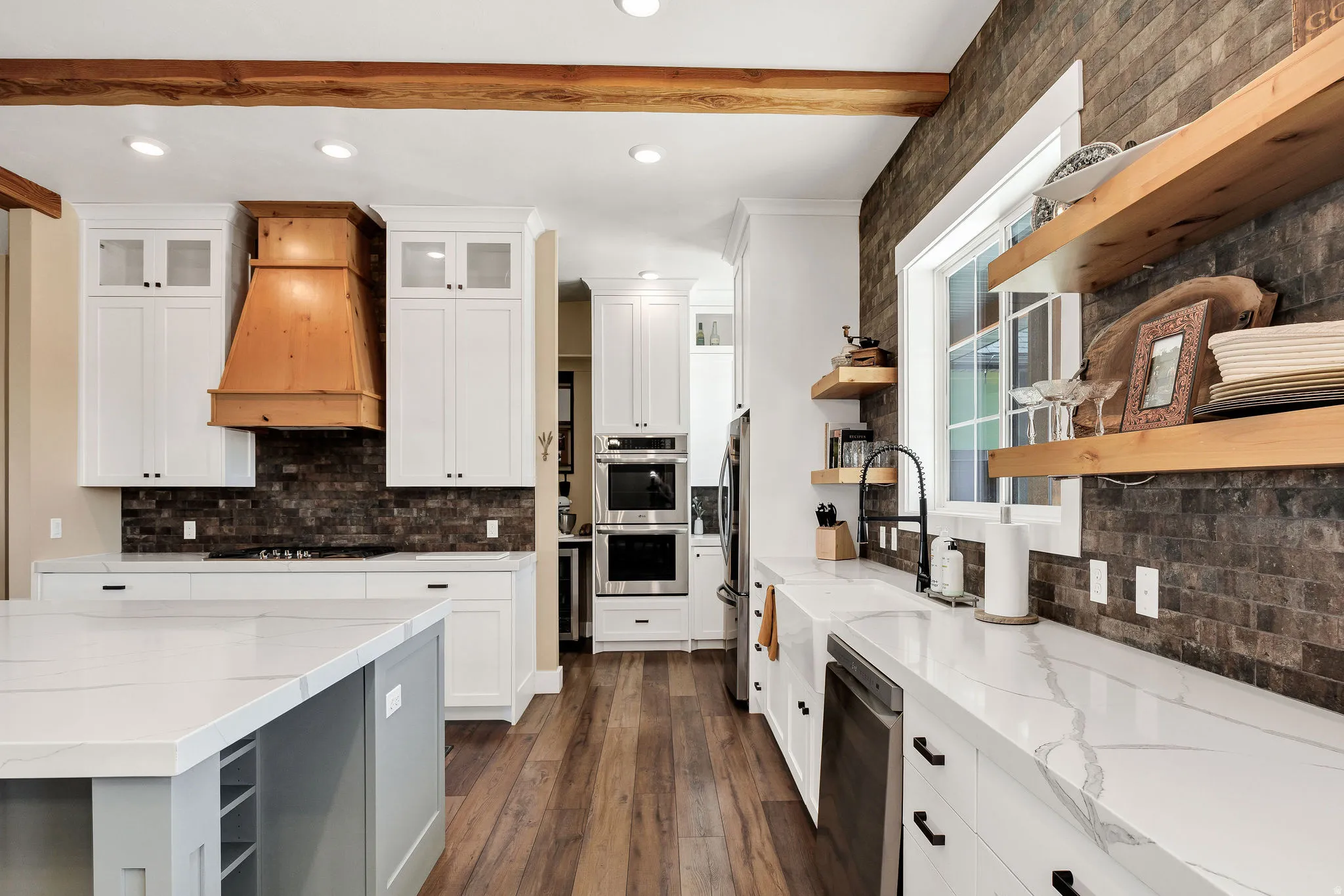 White cabinetry, wood-finish flooring, white countertops, stainless steel double wall ovens, and a brick-finish accent wall with wood shelving