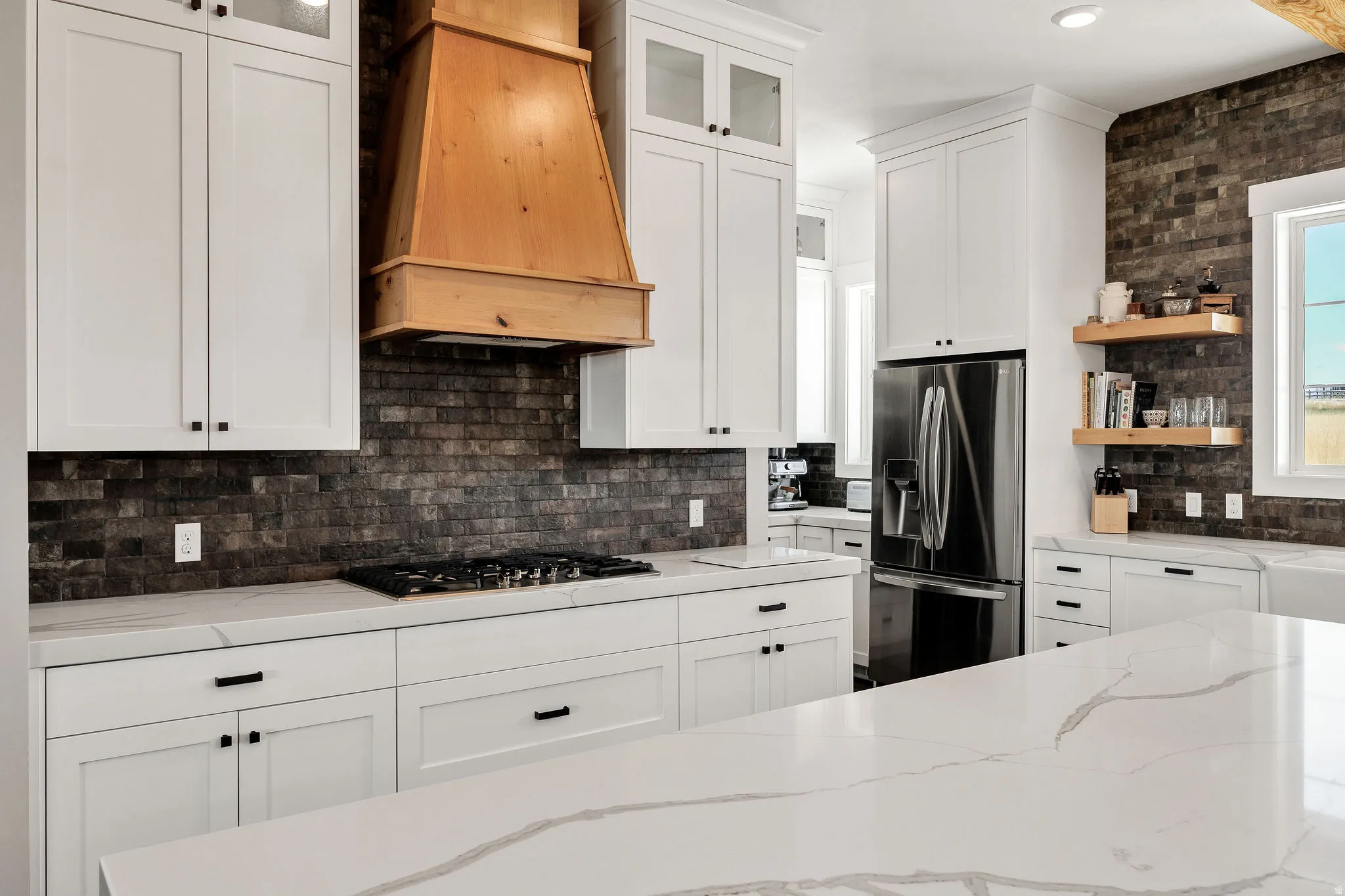 White shaker cabinetry, wood range hood, dark brick-patterned backsplash, stainless steel refrigerator, and light-toned countertops