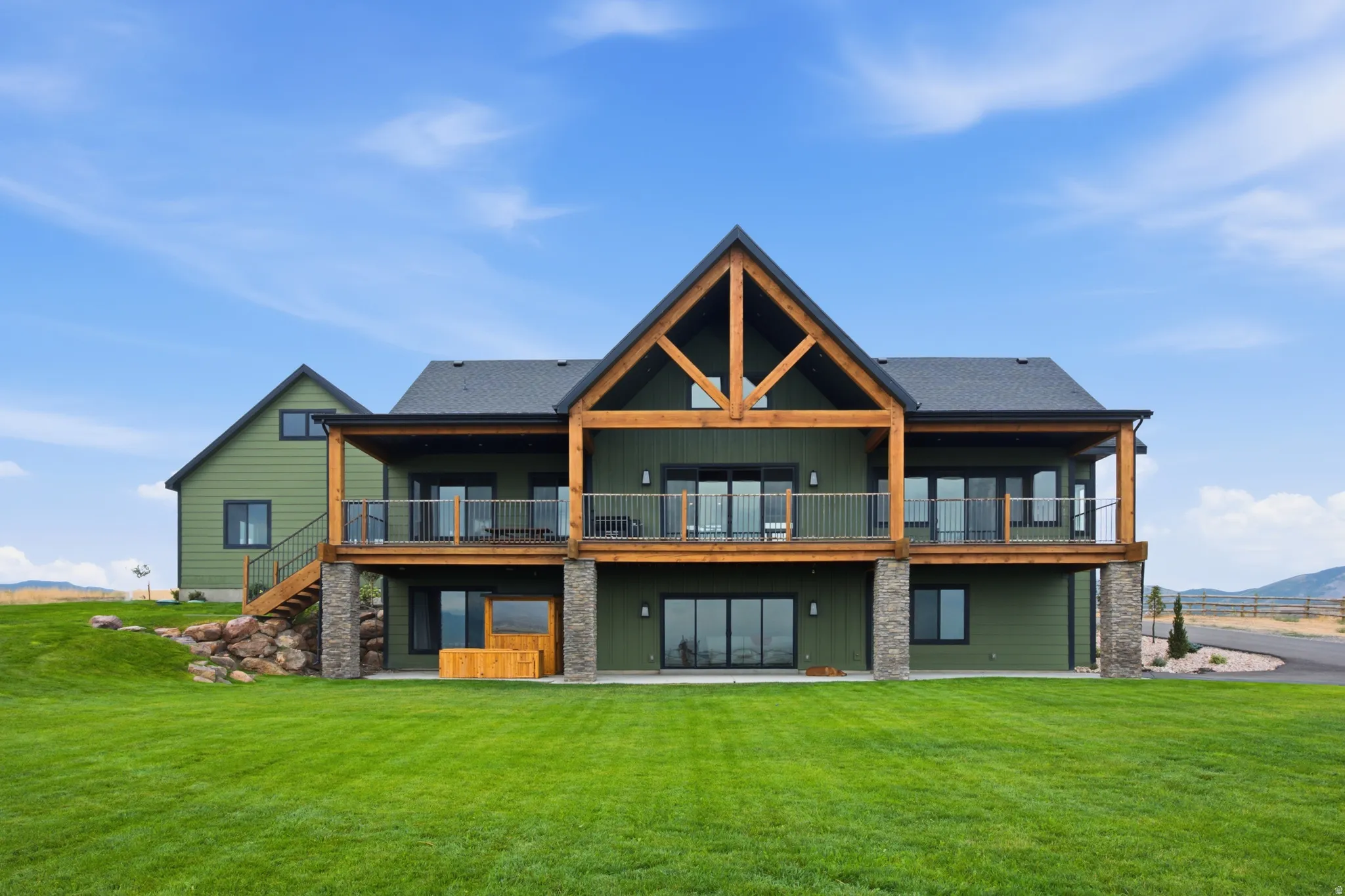 Expansive green lawn with a two-story residence featuring a covered deck, substantial timber frame elements, and stone-clad structural supports