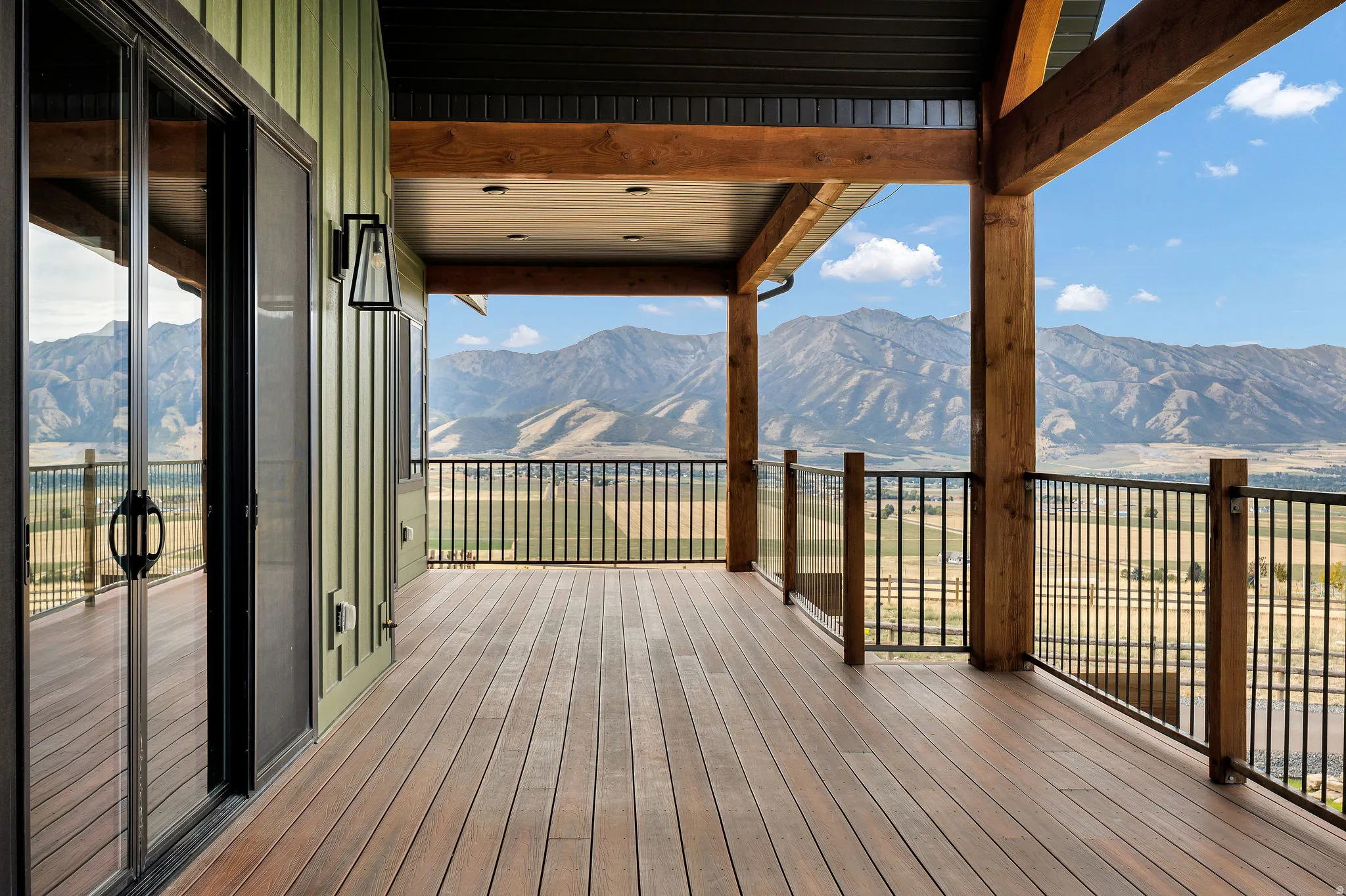 Expansive outdoor deck with wood-finish flooring, black metal and glass panel railing, exposed timber beams, and sliding glass doors