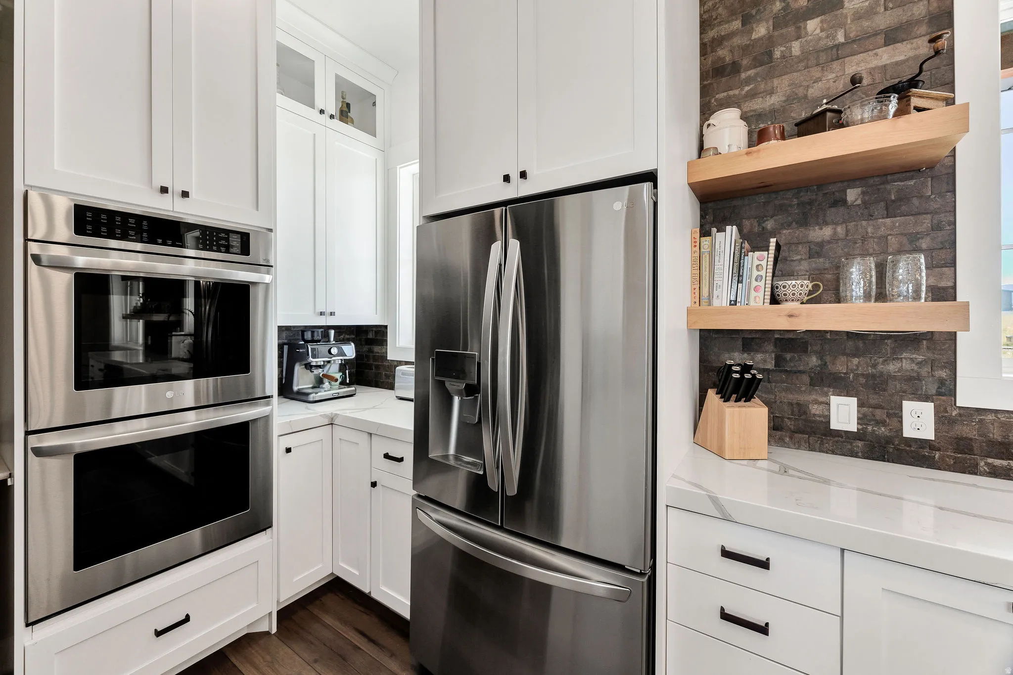 White cabinetry with black hardware, stainless steel double ovens, stainless steel refrigerator, white countertops, and wood-finish flooring