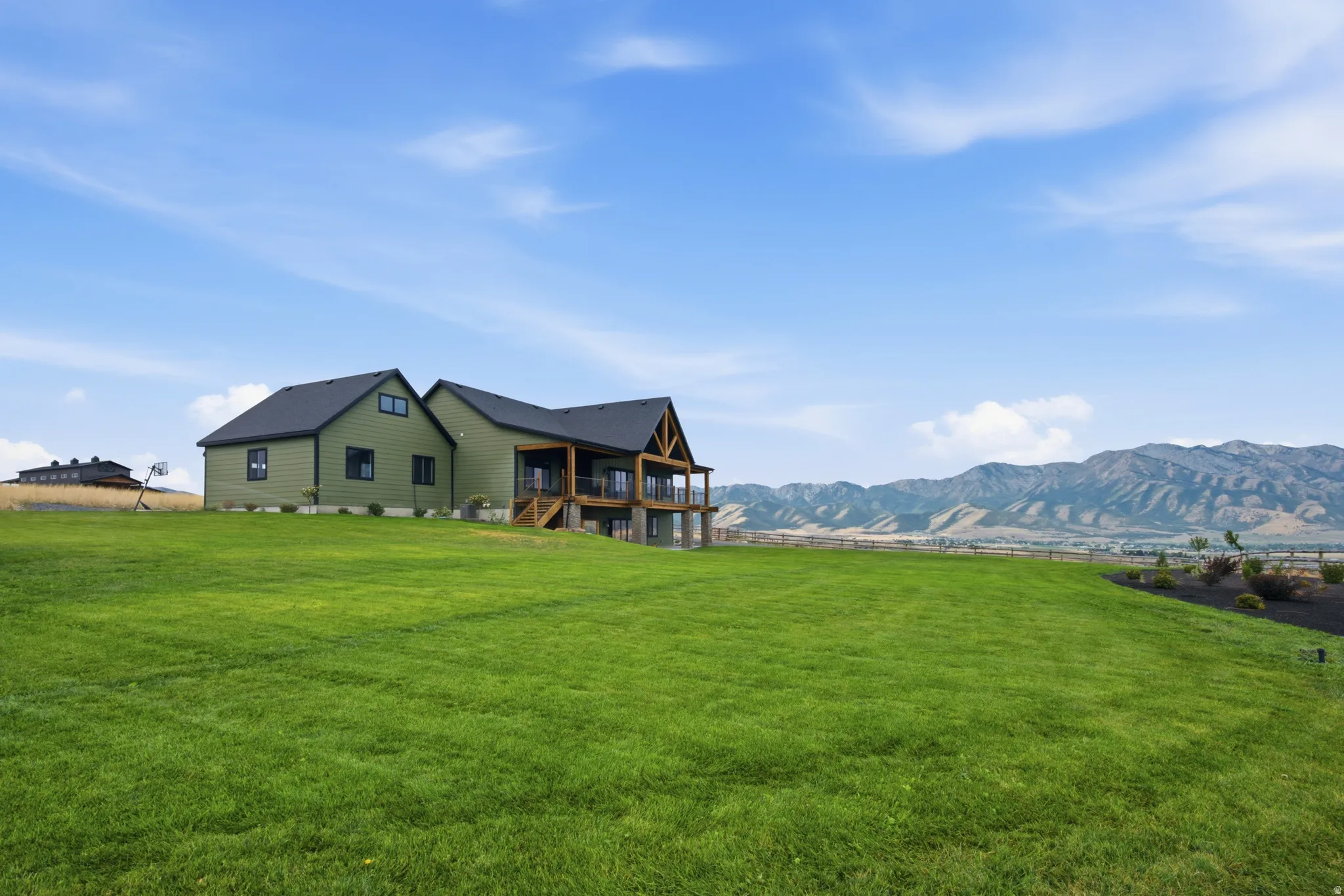 Expansive green lawn surrounding a residence with a covered deck, dark siding, and a prominent mountain range backdrop