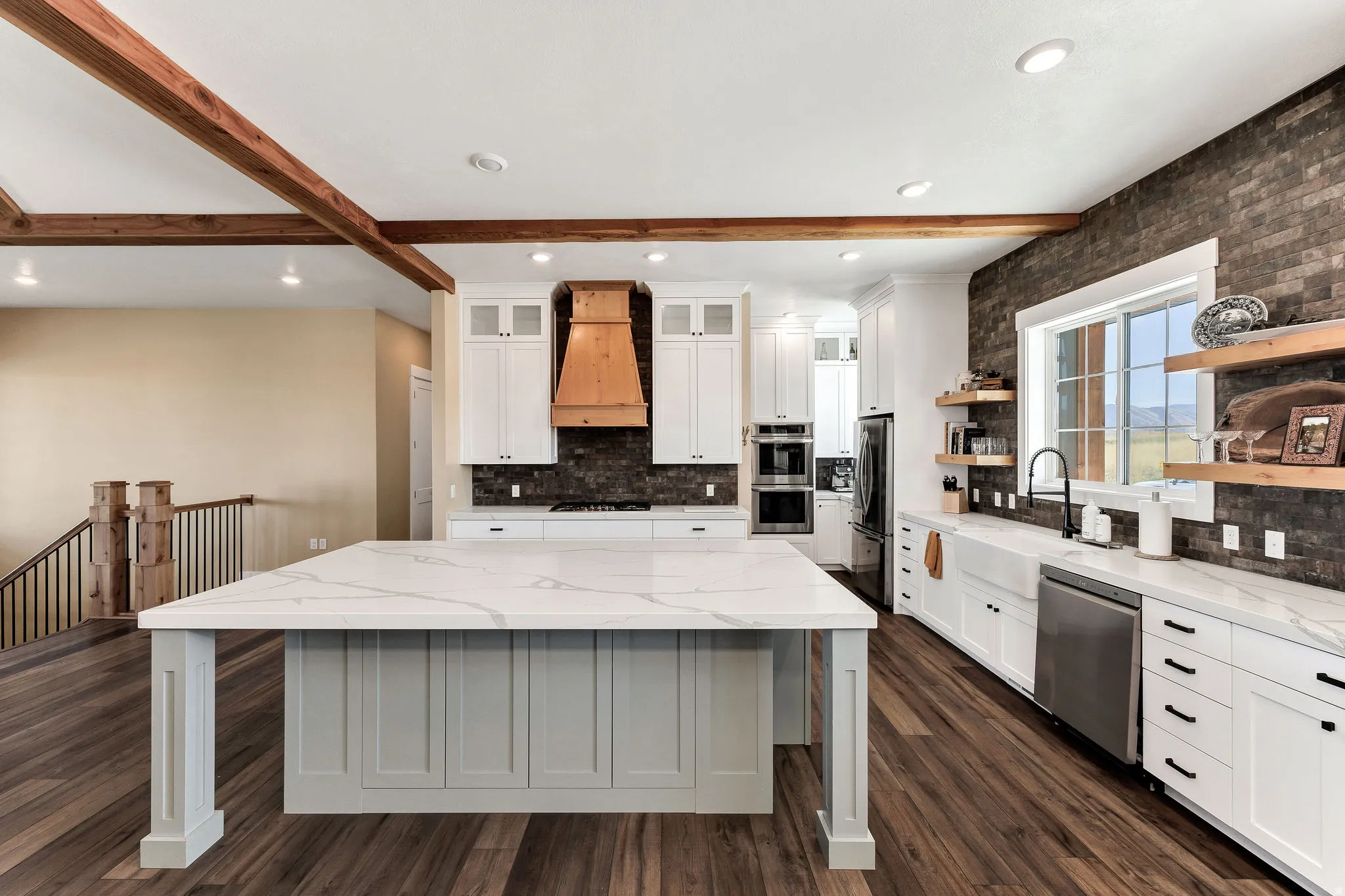 Spacious kitchen featuring a large island with a light-toned countertop, wood-finish flooring, white cabinetry, a wood-paneled range hood, and a dark brick-style accent wall