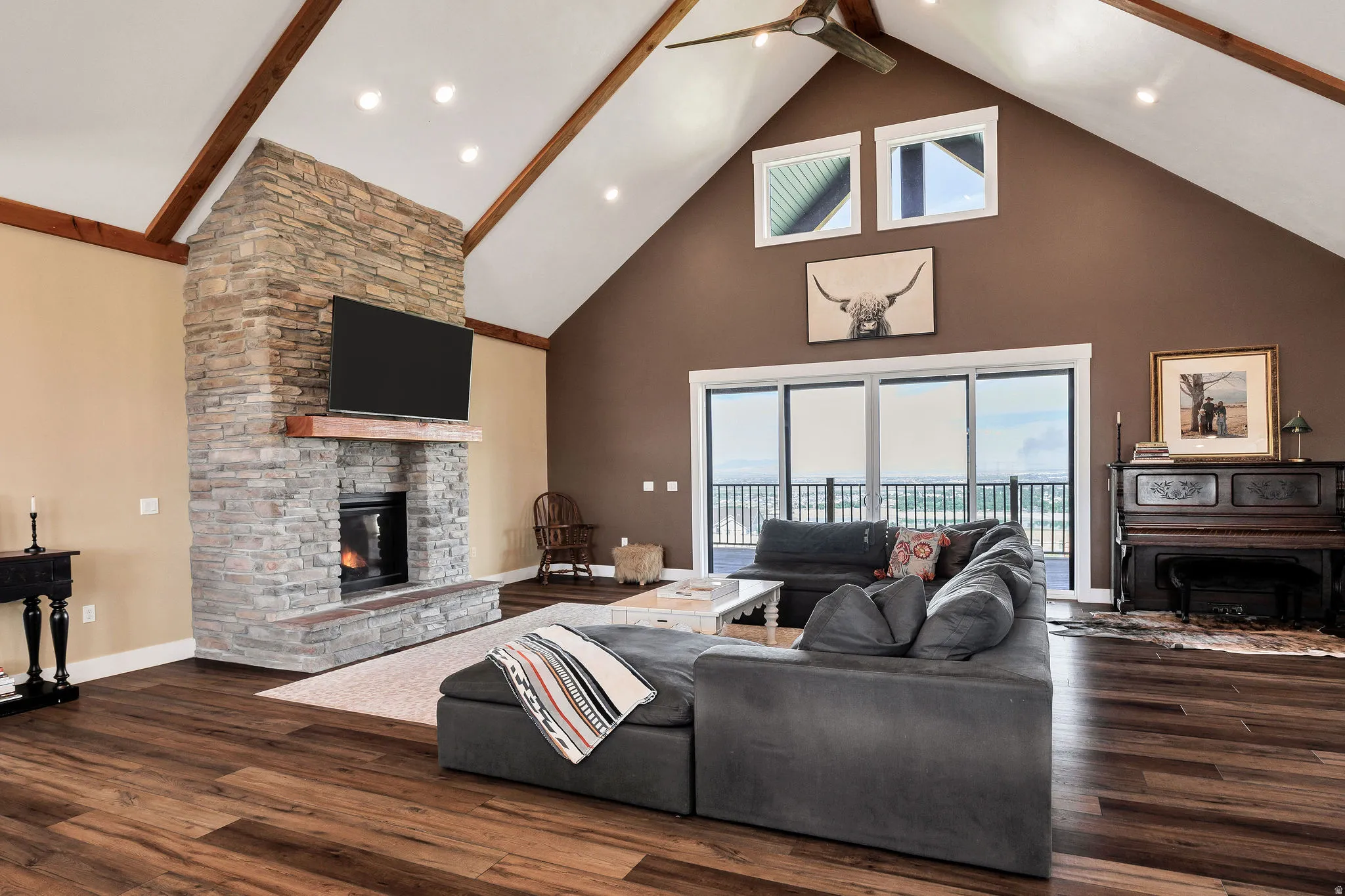 Vaulted ceiling with exposed wood beams, stone fireplace, wood-finish flooring, recessed lighting, and expansive sliding glass doors