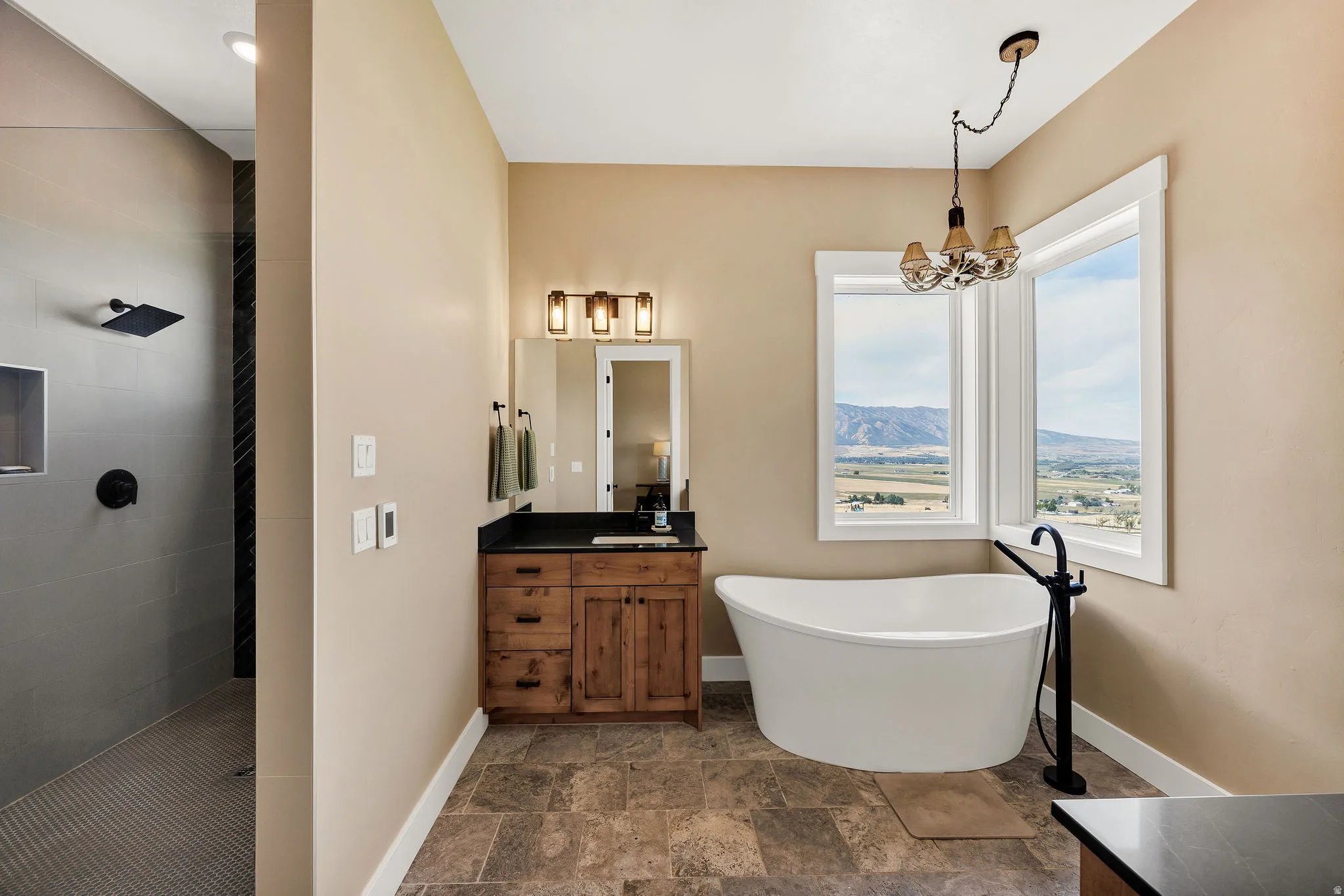 Freestanding soaking tub with floor-mounted faucet, walk-in shower with rainfall showerhead, wood vanity with dark countertop, and tiled flooring