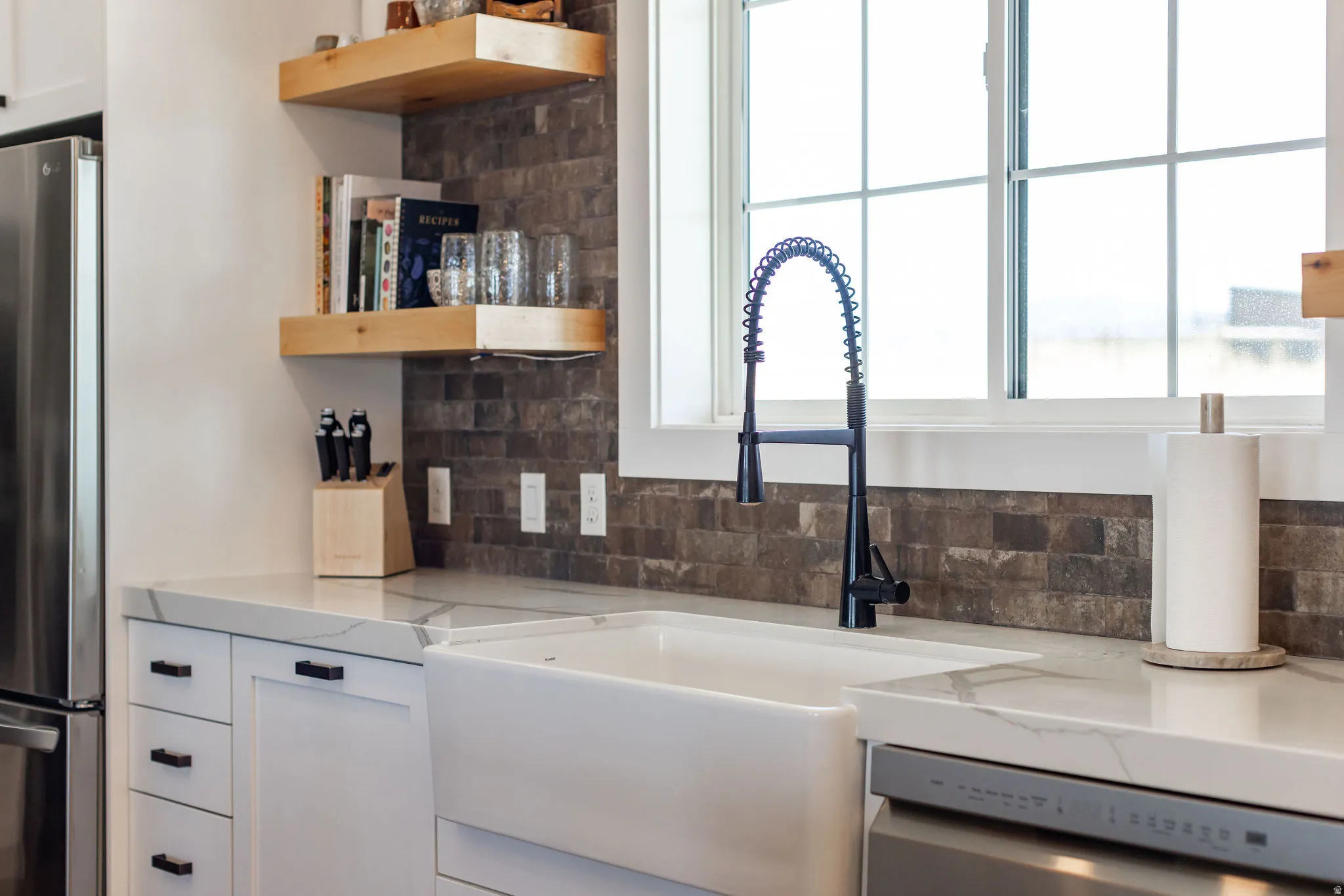 White apron-front sink, quartz countertops, dark tiled backsplash, white cabinetry with dark hardware, and a black gooseneck faucet