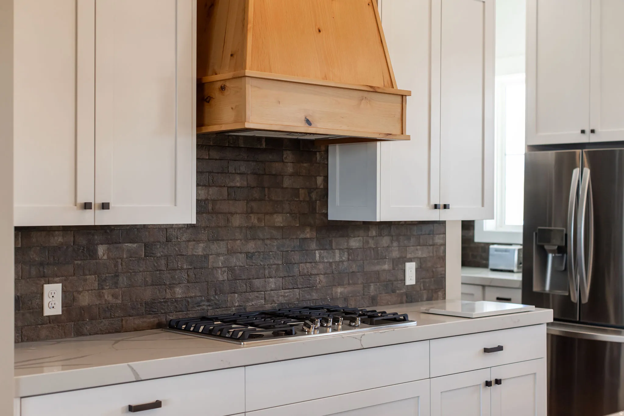White cabinetry with black hardware, wood-finish range hood, dark brick-patterned backsplash, gas cooktop, and light-toned countertop