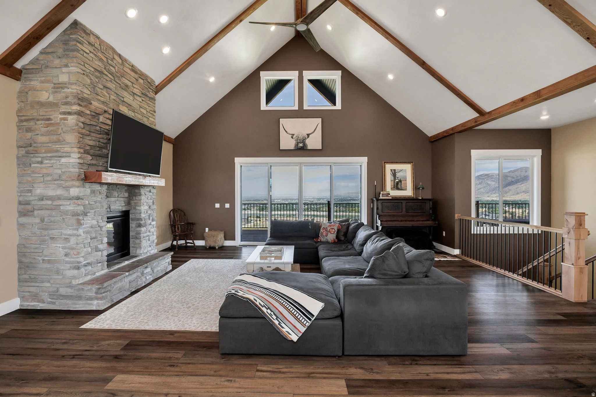 Vaulted ceiling with exposed wood beams, stone-clad fireplace with hearth, wood-finish flooring, and sliding glass doors to a balcony