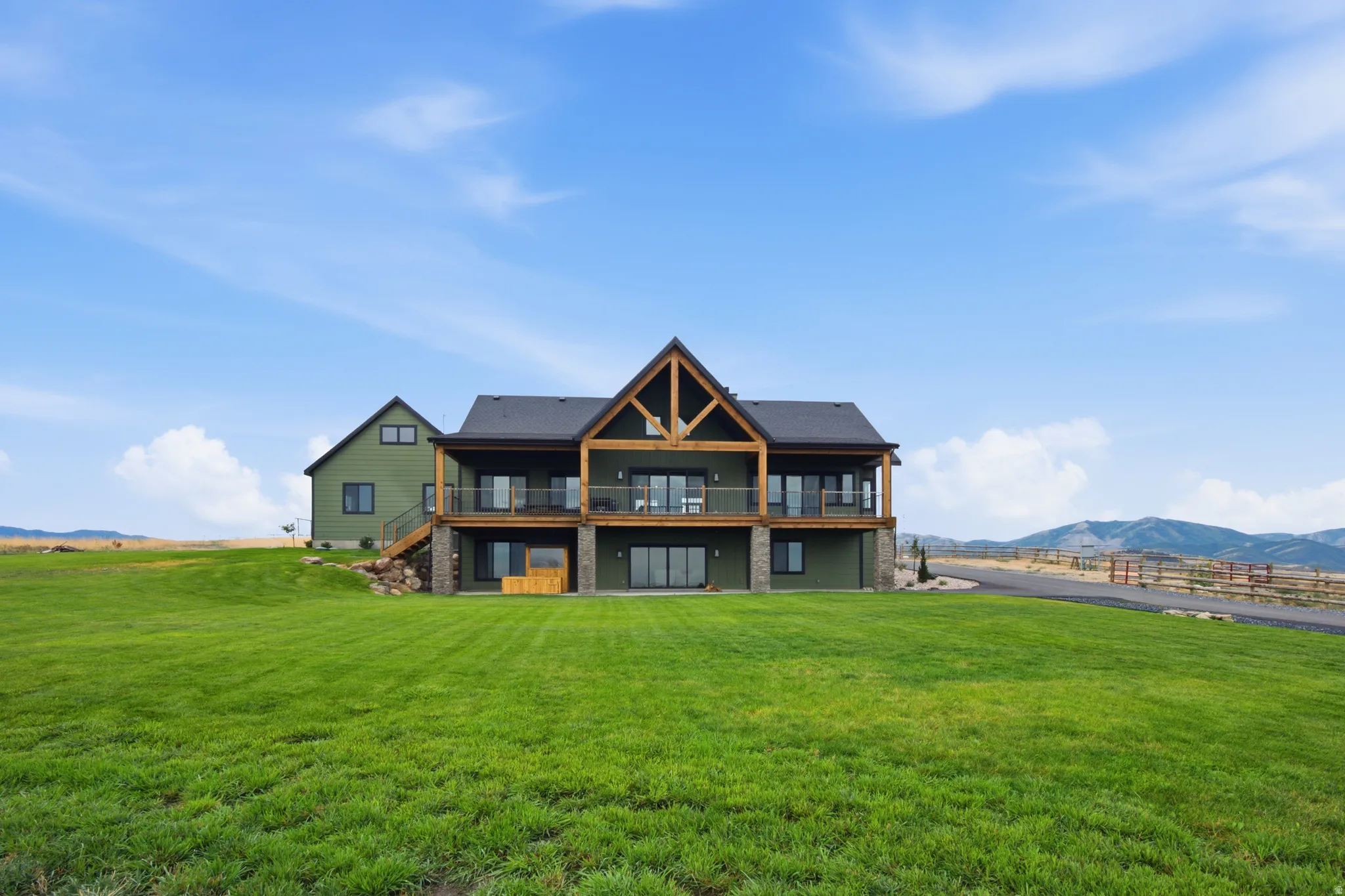 Expansive green lawn surrounds a two-story residence featuring a covered upper-level balcony, a lower-level patio, a prominent gable with exposed wood beams, and a stone-clad foundation