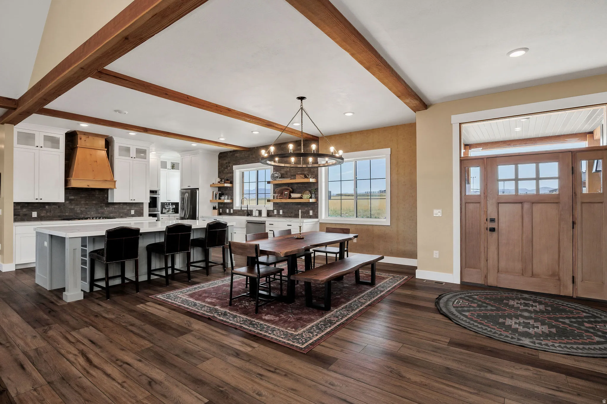 Wood-finish flooring throughout, a kitchen island with seating, exposed ceiling beams, a wood-paneled front door with side panels, and white cabinetry