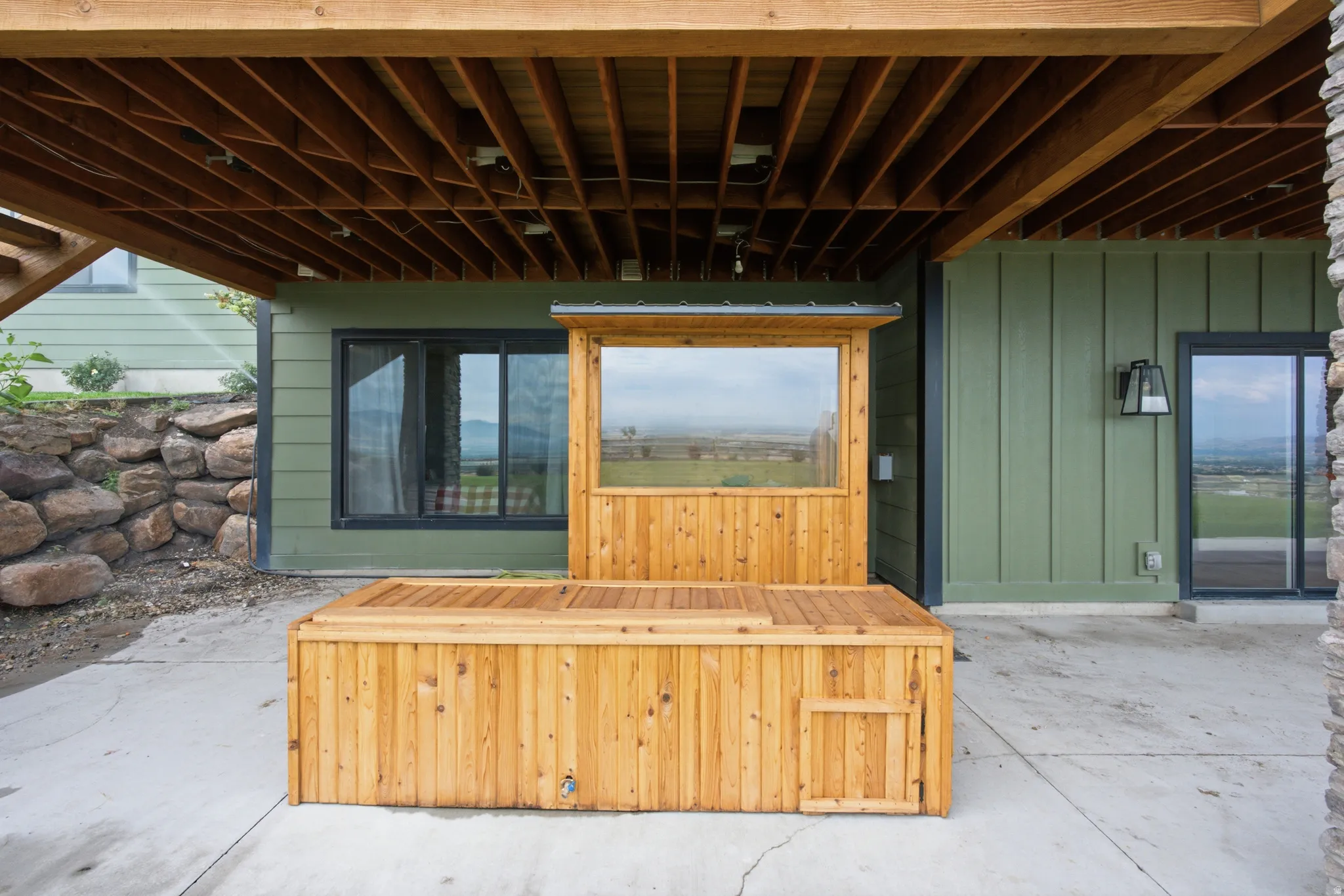 Wood-finished hot tub with a built-in privacy screen, covered concrete patio, exposed wood beam ceiling, green vertical siding, and a stone retaining wall