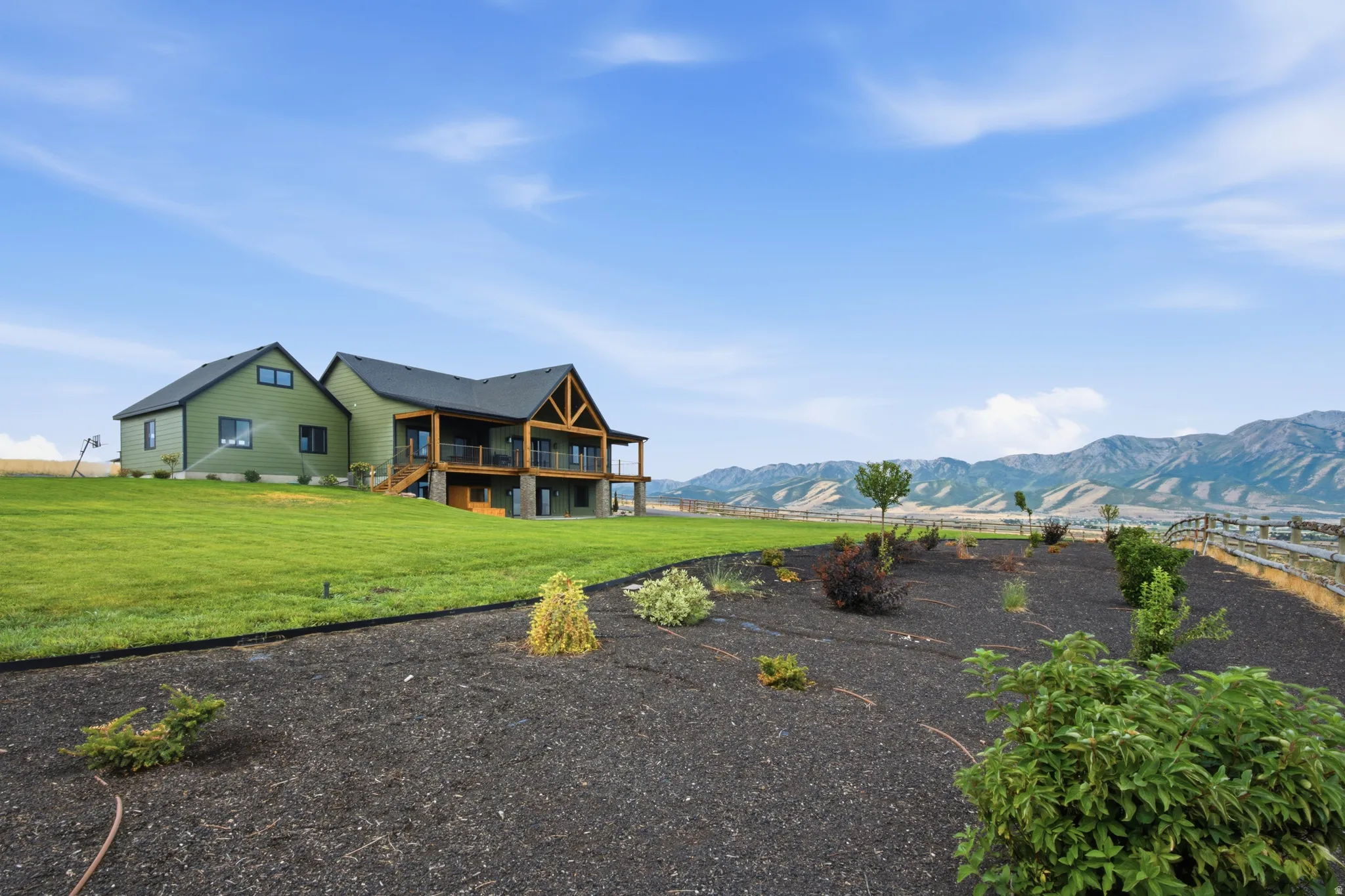 Green façade with dark trim, expansive lawn, two-story deck with wood beams, mountain backdrop, and landscaped planting beds