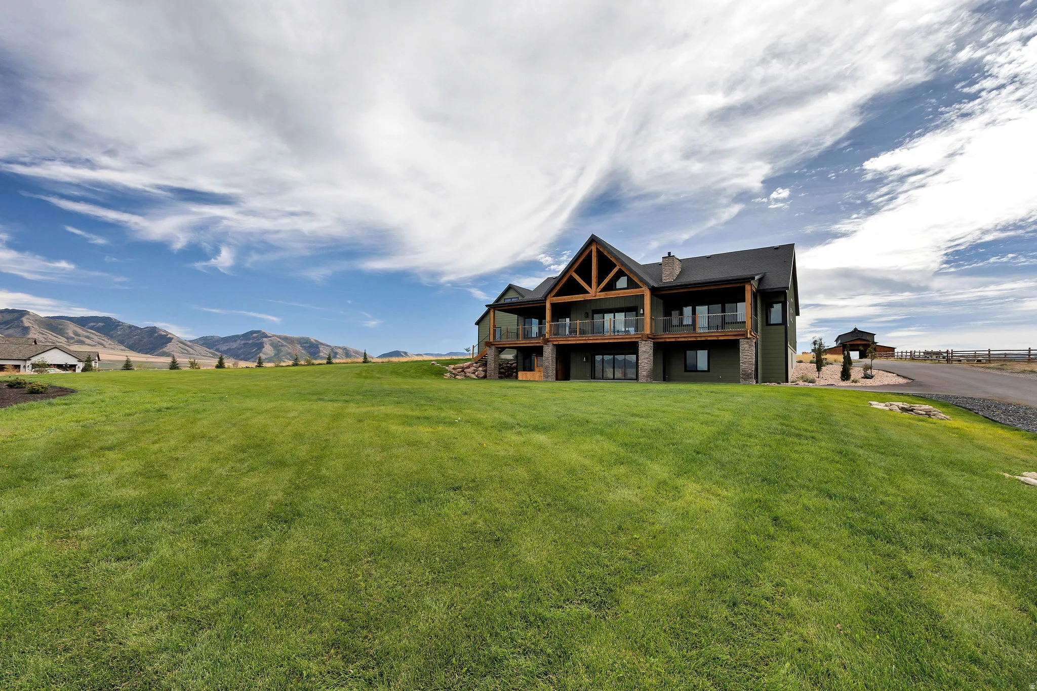 Expansive green lawn surrounds this property featuring a multi-level wood-clad exterior, exposed architectural beams, multiple balconies with glass railings, and a paved driveway leading to a separate structure