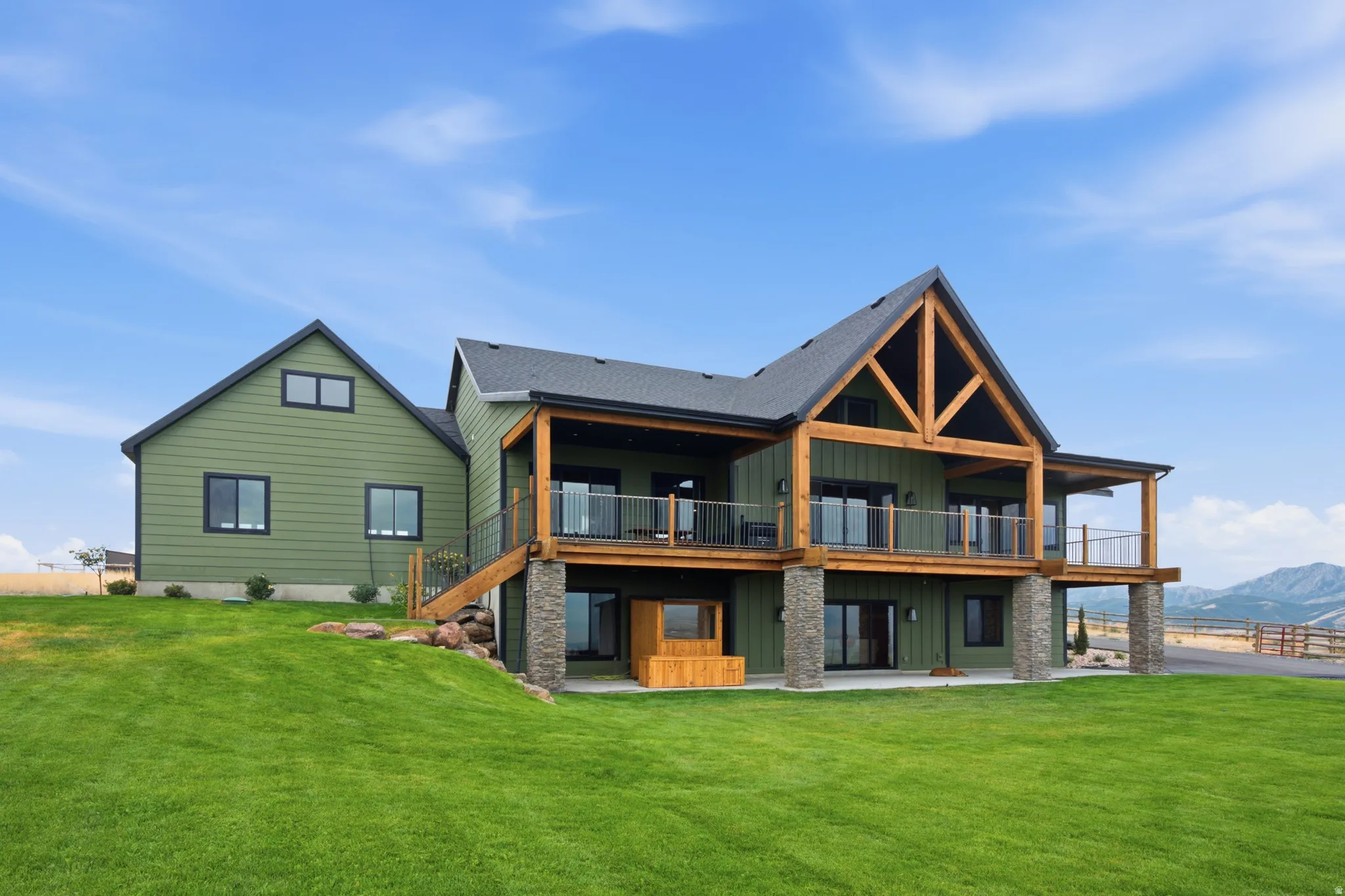 Expansive green siding exterior with a pitched roof, an upper-level balcony with wood beams and metal railings, a lower-level patio, and stone pillar accents
