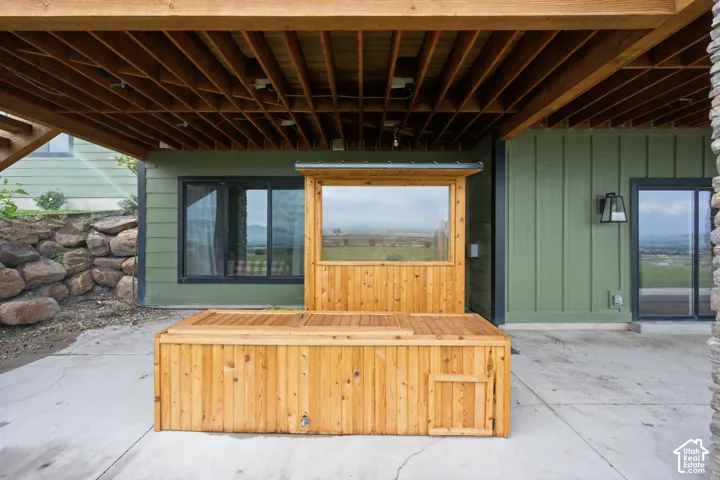 Wood-finished hot tub with a built-in privacy screen, covered concrete patio, exposed wood beam ceiling, green vertical siding, and a stone retaining wall