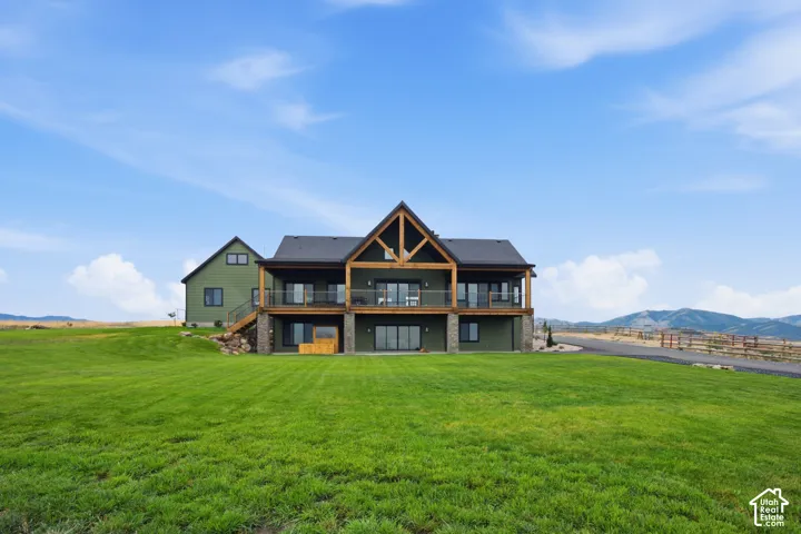 Expansive green lawn surrounds a two-story residence featuring a covered upper-level balcony, a lower-level patio, a prominent gable with exposed wood beams, and a stone-clad foundation