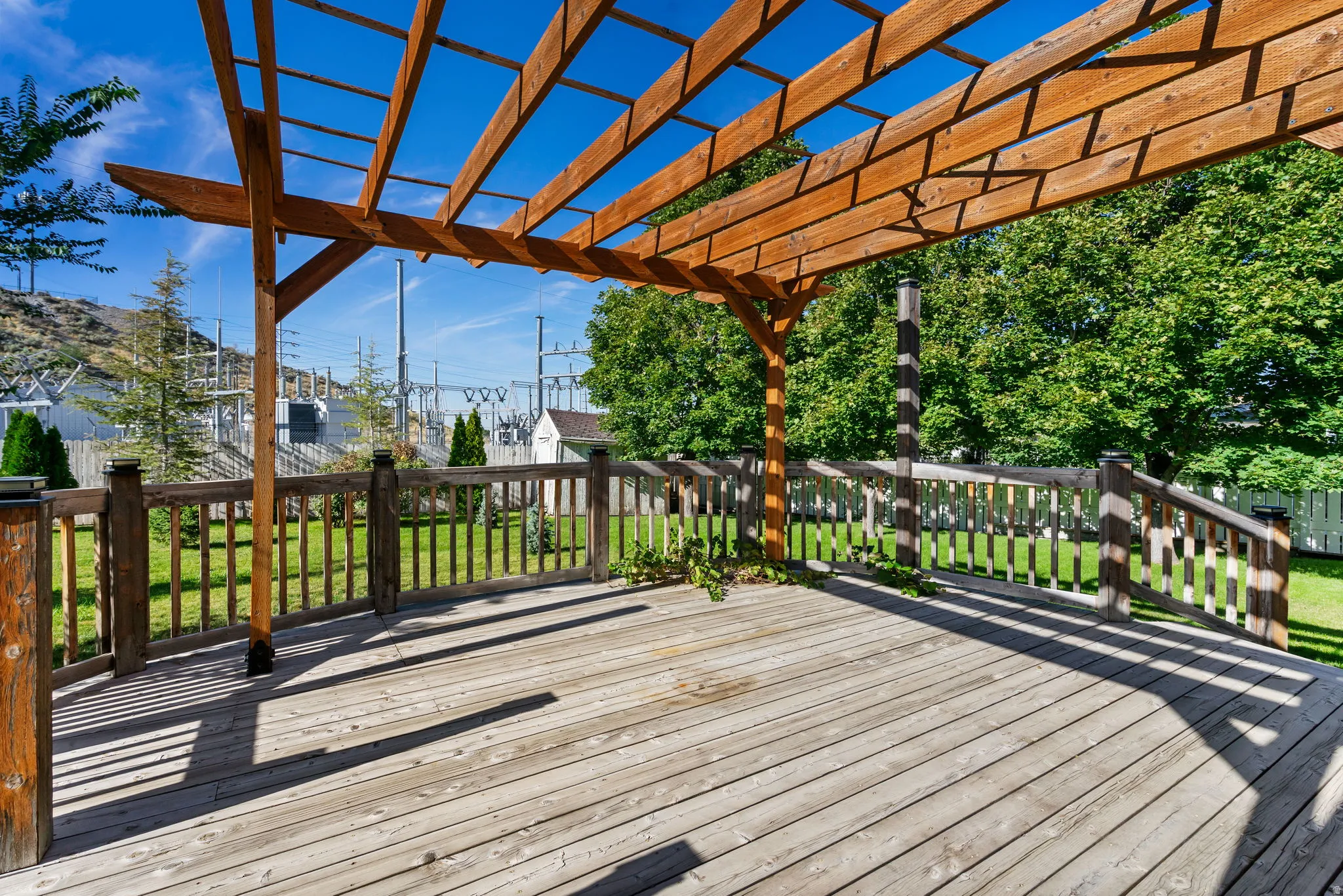 Expansive wood deck with a slatted pergola, surrounded by a wood railing, overlooking a green lawn with mature trees