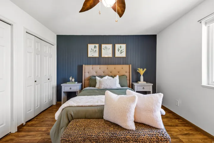 Bedroom featuring wood-finish flooring, a decorative accent wall, a ceiling fan with light fixture, and a window with vertical blinds