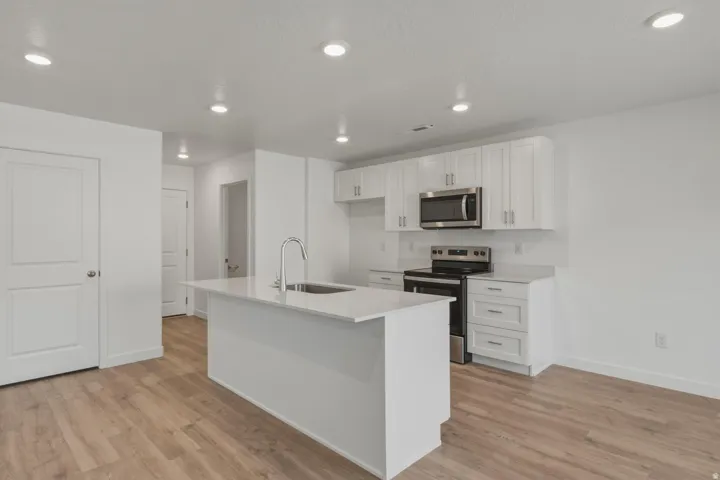 Kitchen featuring a central island with an integrated sink, white cabinetry, stainless steel appliances, and wood-finish flooring