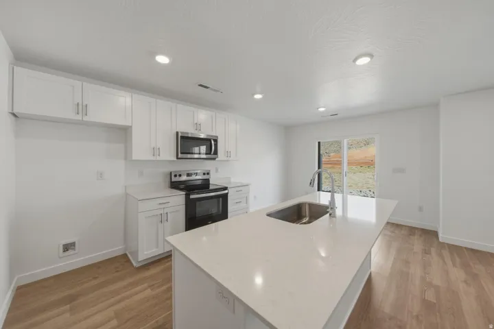 Kitchen featuring an island with an integrated sink, white cabinetry, stainless steel appliances, light countertops, and wood-finish flooring