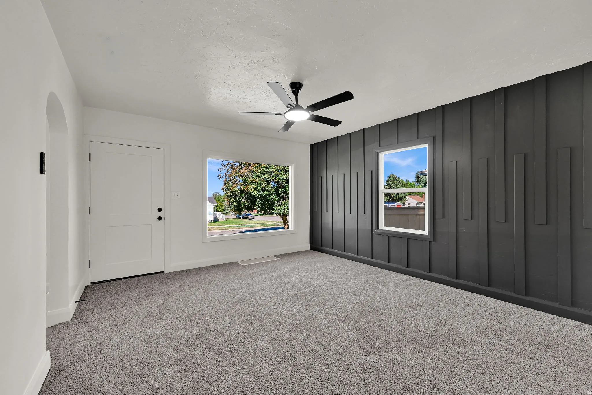 Archway entry, white paneled door, large picture window, textured accent wall, and ceiling fan with integrated lighting