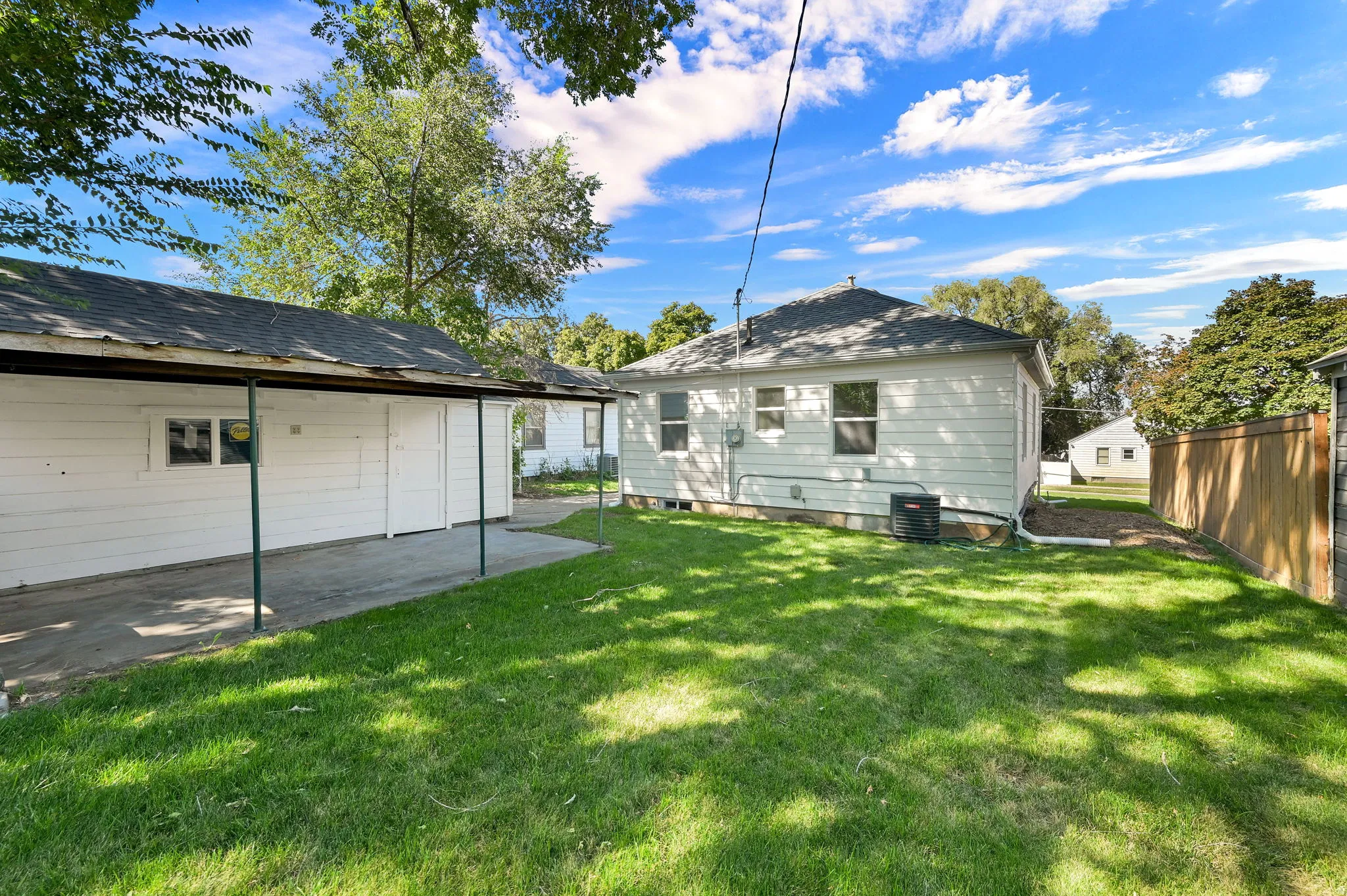Expansive rear yard featuring green lawn, established trees, and a carport with a concrete driveway
