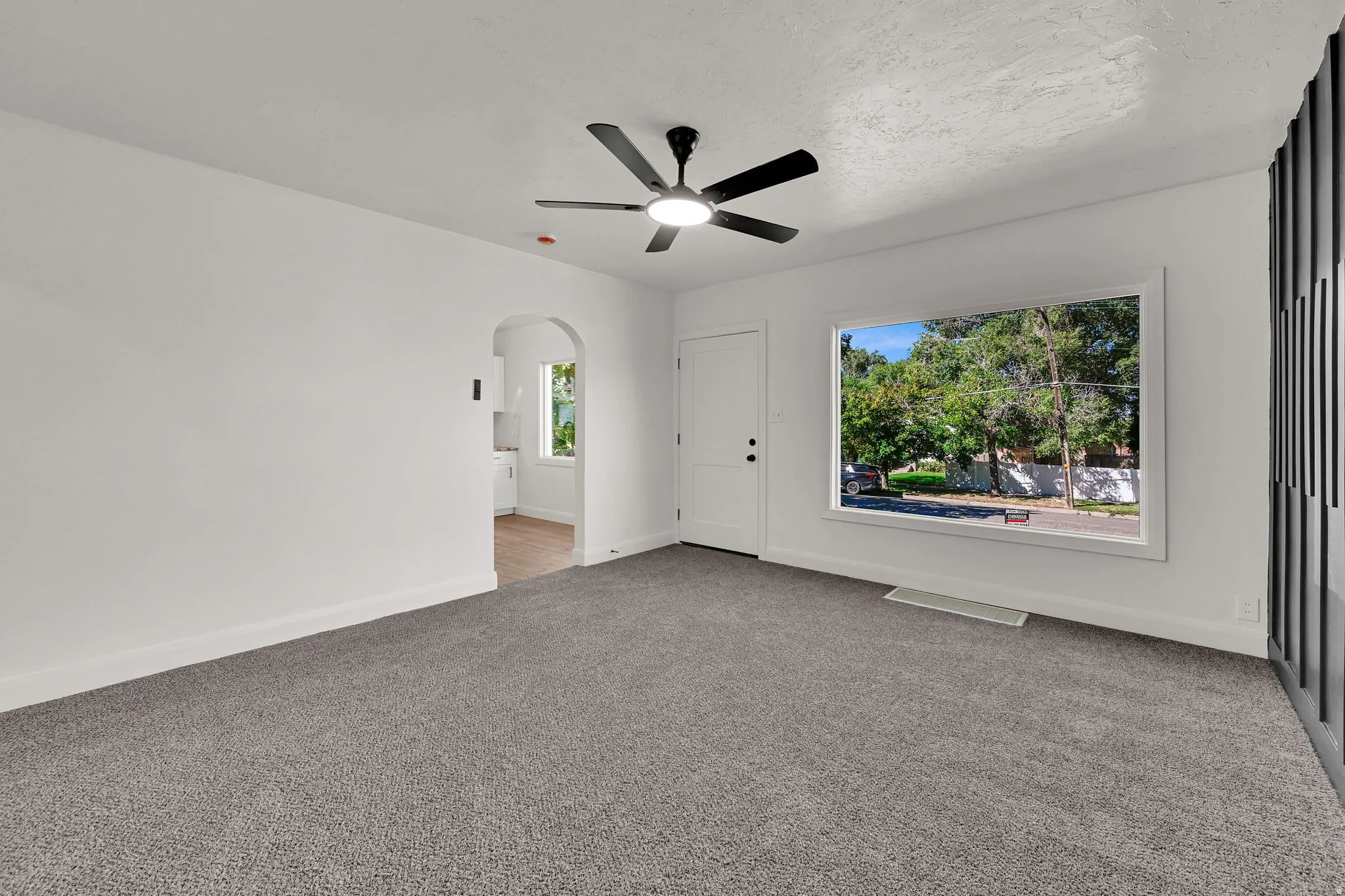 Newly installed grey carpeting, modern ceiling fan with integrated lighting, arched doorway, and a white entry door