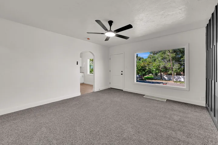 Newly installed grey carpeting, modern ceiling fan with integrated lighting, arched doorway, and a white entry door