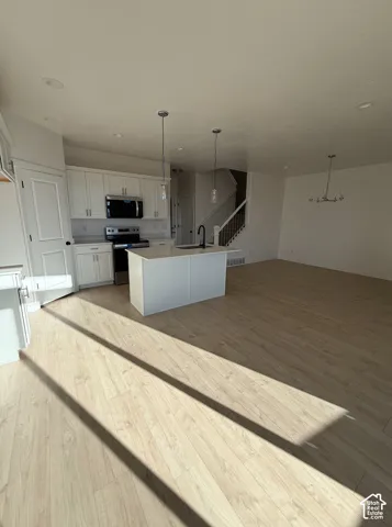 Open-concept living space featuring wood-finish flooring throughout, a white cabinetry kitchen with black appliances and an island with countertop seating, pendant lighting, and a grand staircase with wrought iron railings