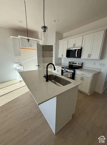 Kitchen featuring white cabinetry, light-toned wood-finish flooring, a center island with a stainless steel sink, light-colored countertops, and stainless steel appliances