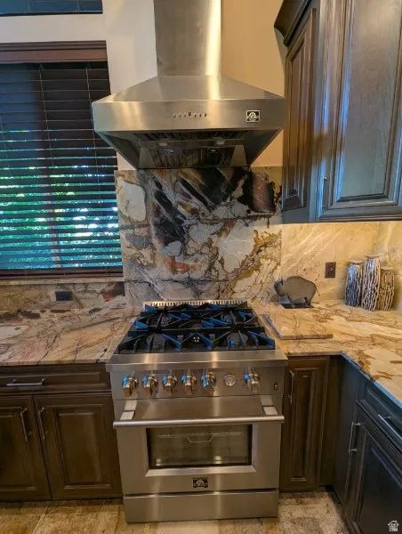 Stainless steel range with a professional-grade hood, natural stone countertops, a full-height natural stone backsplash, and dark wood-finish cabinetry