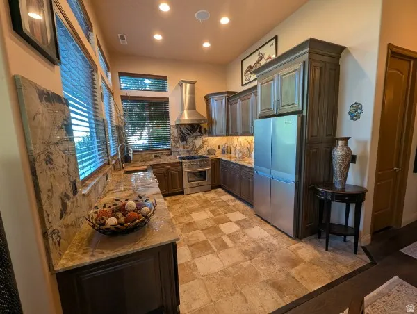 Kitchen featuring stainless steel appliances, dark wood cabinetry, light-toned stone countertops, a matching stone backsplash, and recessed lighting