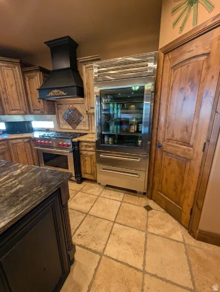 Built-in Sub Zero stainless steel refrigerator with glass panel, custom range hood, wood-finish cabinetry, stone countertop, and tile flooring