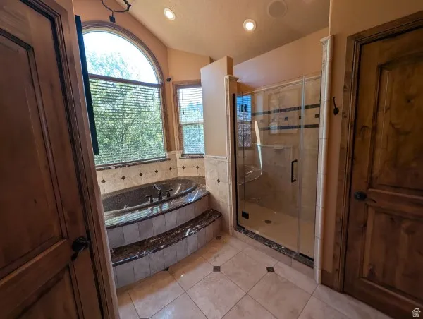 Primary bath with tiled flooring, built-in soaking tub with tiled surround, arched window, walk-in shower with glass enclosure, and wood-panel doors