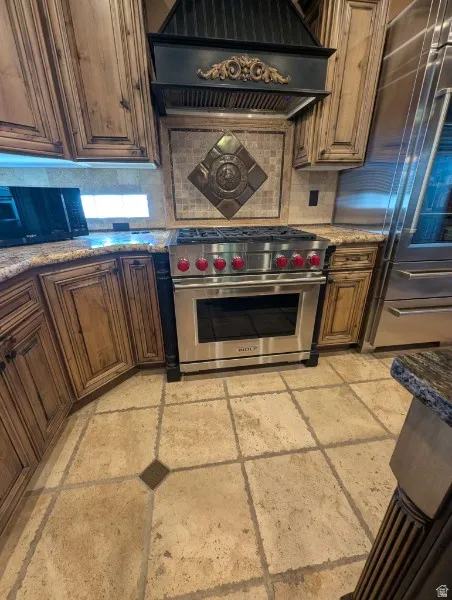 Chef's kitchen featuring professional-grade Wolf stainless steel & electric range with multiple burners, custom wood cabinetry with decorative glazing, coordinating stone countertops, decorative tile backsplash with diamond inlay, black range hood with or