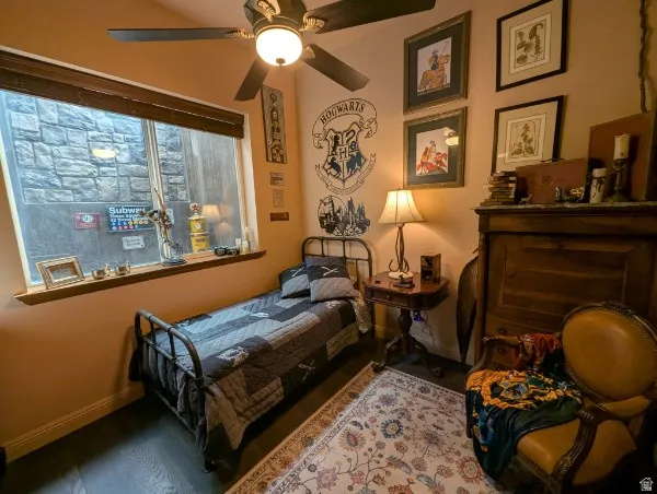 Recessed window with wood blinds, dark wood-finish flooring, ceiling fan with integrated lighting, and a built-in wood-paneled feature in basement bedroom