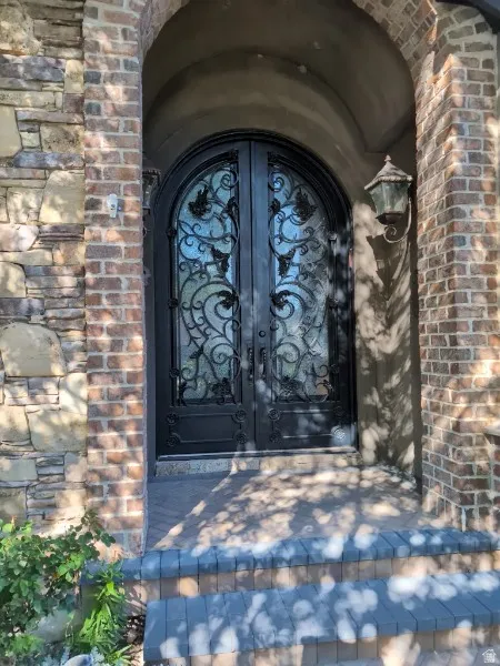 Arched entry featuring a wrought iron double door with decorative glass, brick and stone facade, exterior wall-mounted lantern, and paver steps