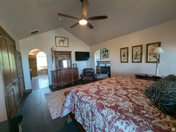Primary bedroom with vaulted ceiling with a ceiling fan, dark wood-finish flooring, two arched doorways, and a built-in fireplace in an adjacent area