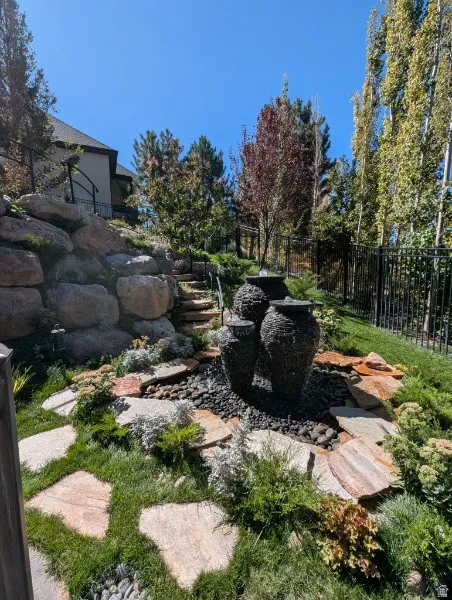 Tiered water feature with a stone rock bed, stone steps, landscaped garden with varied plantings, and a black wrought iron fence
